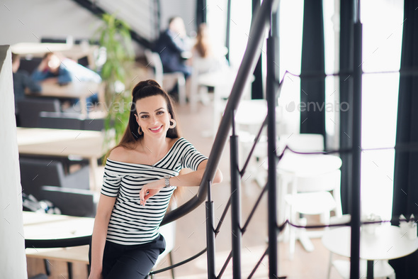 Attractive smiling woman with natural makeup is standing in a cafe and looking at camera. Stock Photo by romankosolapov
