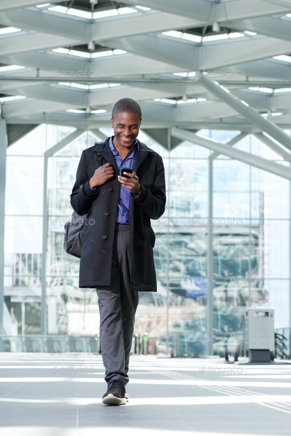 smiling black businessman walking with cell phone at airport Stock Photo by mimagephotography