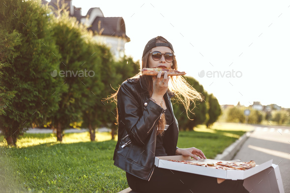 Cheerful young woman enjoying slice of pizza while sitting on curb outdoor Stock Photo by Vladdeep