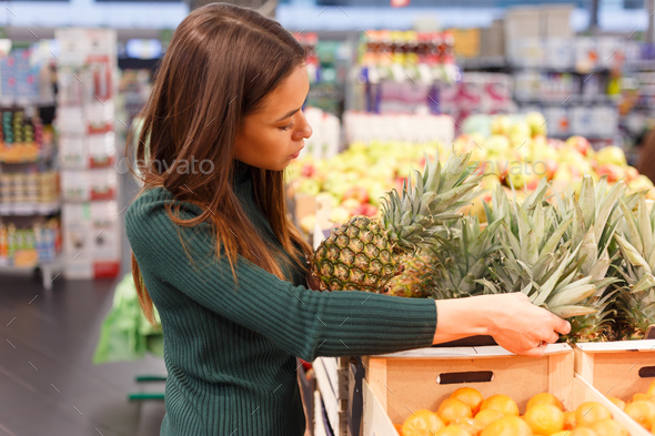 Young brunette woman buying pineapple at grocery shop. Stock Photo by Vladdeep