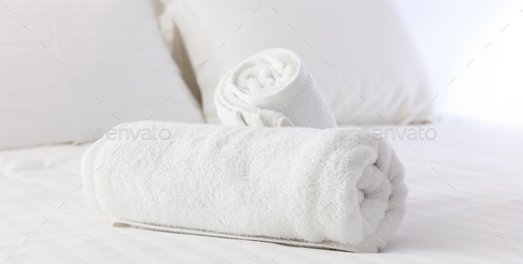 Hotel's bedroom. White fluffy, rolled towels, linen sheets and pillows on a bed. Close up view. Stock Photo by rawf8