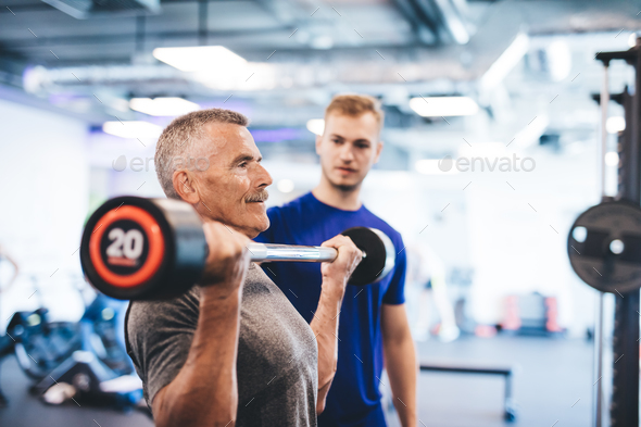 Older man lifting weights, supervised by gym assistant. Stock Photo by photocreo