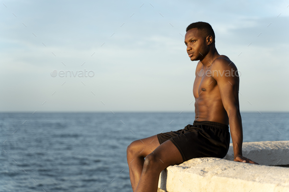 Man With Naked Torso Standing On The Beach And Looking Away While Warming At The Sun Stock Photo