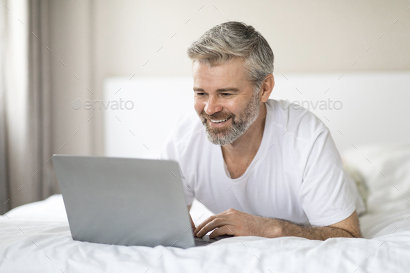 Relaxed Mature Man Lying On Bed Using Laptop Copy Space Stock Photo