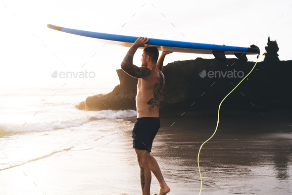 Shirtless Man Walking On Sandy Beach With Surfboard Above Head Stock