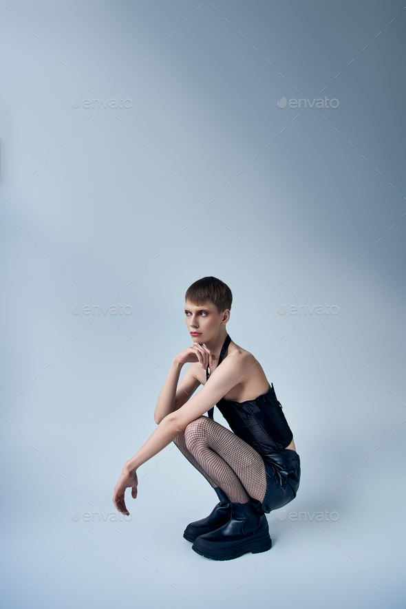 Queer Model In Black Corset And Fishnet Tights Sitting On Grey Backdrop