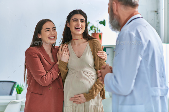 Joyous Lesbian Couple With Hands On Pregnant Belly Smiling At Their Gynecologist Ivf Concept