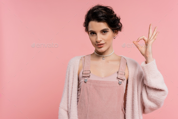 Smiling Brunette Woman In Strap Dress And Fluffy Cardigan Showing Okay Sign Isolated On Pink