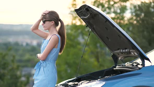 Young woman driver standing near a broken car with popped up hood waiting for help to arrive. alt