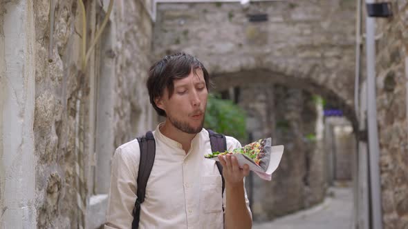 A Young Man Eats a Delicious Pizza on Black Bread Standing on a Street of an Old European Town alt
