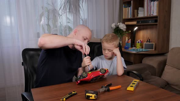 A Man and a Little Boy are Sitting at a Table and Repairing a Broken Toy Car alt