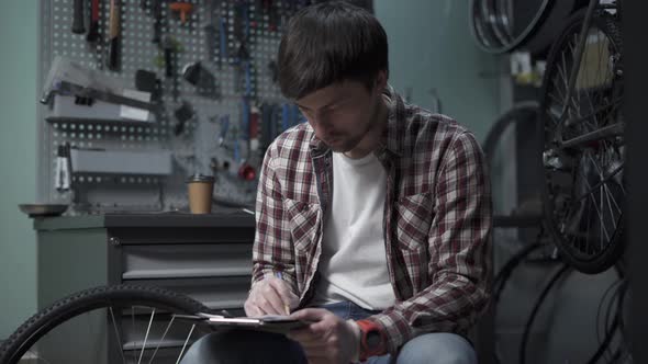 A Male Bicycle Mechanic Calculates the Cost of Repairing a Cycle with a Pen and Clipboard in Hand alt