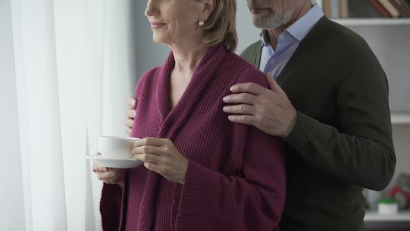 Elderly Female with Cup of Tea by Window, Man Hugging Behind, Kissing on Cheek alt