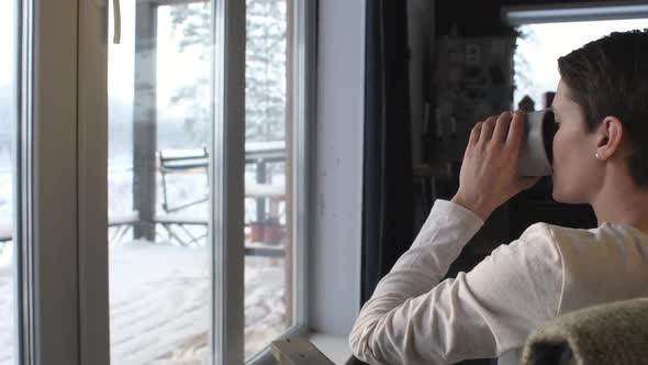 woman sits on  chair window looks at winter landscape and drinks coffee from mug alt