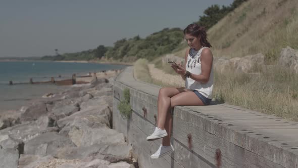 A Young Attractive Woman Sat By The Sea Using Her Phone To Order A Product Online - Ungraded alt