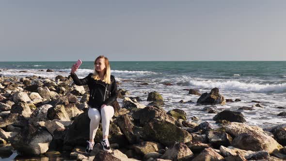 Beautiful Girl Sitting on a Rock By the Sea and Posing for the Camera Taking a Selfie alt