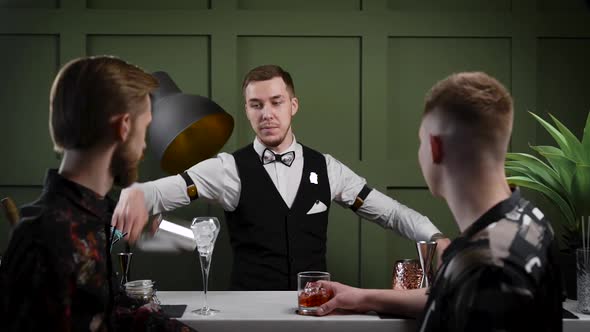 A Male Bartender Prepares Cocktails. Two Friends of the Visitors Are Waiting for Their Drinks at the alt