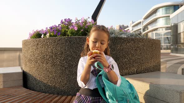 Little Pupil in School Uniform is Eating Apple on Bench