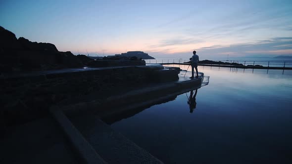 The Profile Of A Man Beside The Swimming Pool Capturing The Sunset In Guernsey. -wide shot alt