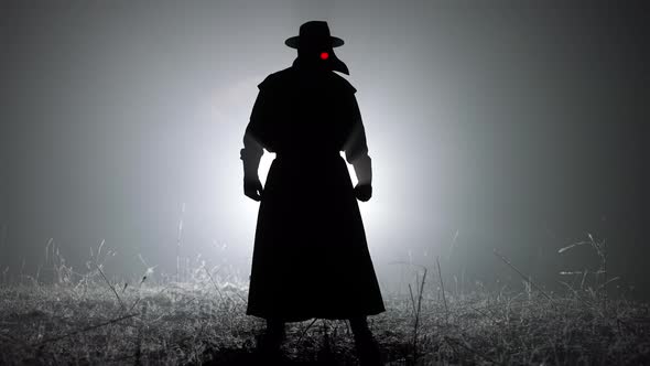 Silhouette of Scary Plague Doctor Man in Long Mantle Costume Standing in Front of Bright Spotlight alt