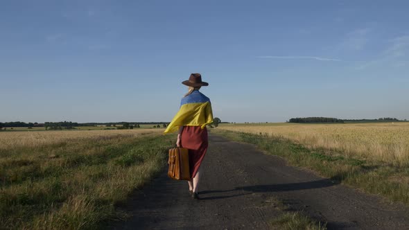 Girl in Ukraine flag with suitcase on country road in sunset