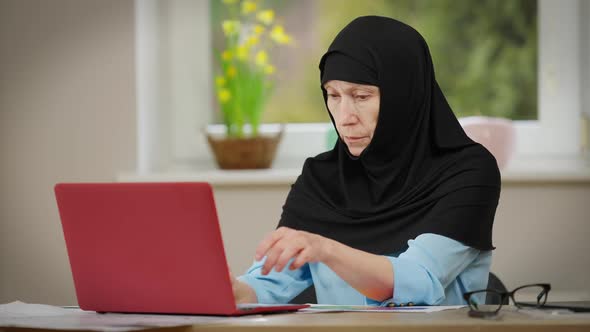 Concentrated Mature Woman in Black Hijab Typing on Laptop Keyboard Sitting at Table Indoors alt