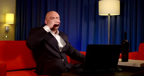 a Bald Man in a Dark Suit Sits on a Red Sofa Against a Blue Curtain. He Talks Cheerfully Over a alt