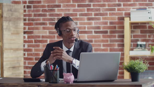 Wearing a Headset and Glasses a Black Man in Suit Speaks Via Video Link Through Laptop at an Office alt