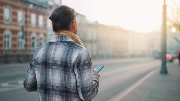 Back View of a Businesswoman in a Coat Walking Around the Old City at the Dawn and Using Smartphone alt