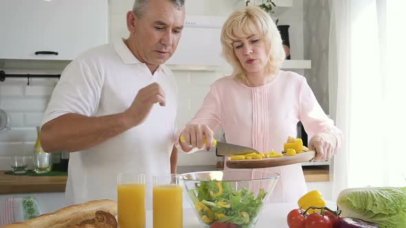 Senior Man Helping Mature Wife Putting Cut Pepper To Salad Bowl alt