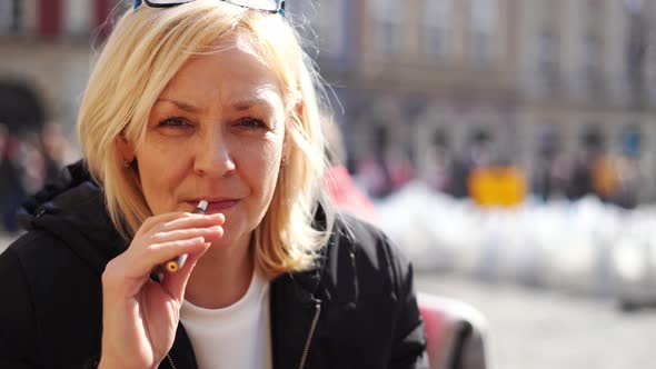 Middle Aged Blonde European Woman Smoking Heated Tabacco Ecigarette Outdoors on the Street alt