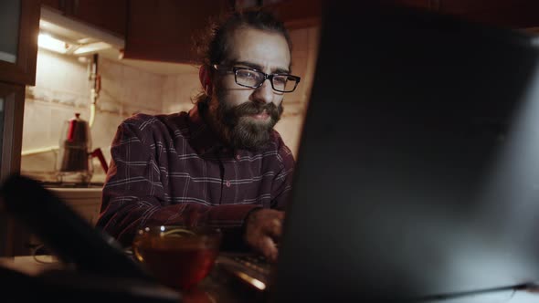 Teenager in Checkered Shirt with Long Beard in Doing Research on Internet from Laptop in Kitchen