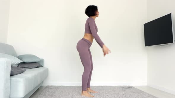 Side view young woman in violet activewear stands on carpet do butt exercises deep squat