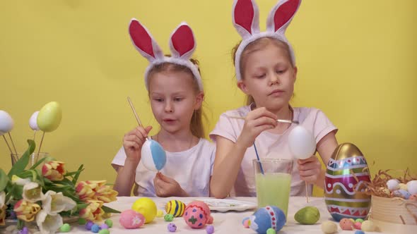 Focused Sisters with Bunny Ears Standing Near Table with Palette and Bouquet of Tulips Coloring Eggs