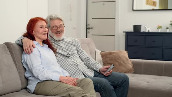 Happy Senior Couple Aged 6070 Watching TV and Laughing alt