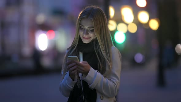 Cute Girl Uses Smartphone and Smiles Standing on Night Street in the City.