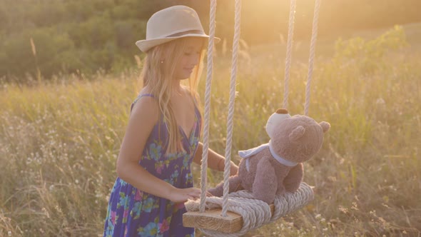 Happy Funny Girl in Hat Is Playing with a Teddy Bear on a Rope Swing During Sunset. Kid Playing on alt