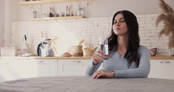 Young Thirsty Woman Holding Glass, Make Sip, Drinking Water, Sitting at Table in Home Kitchen alt