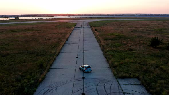 Flying over a car that is traveling on an airplane runway. Forest and grass on the sides. alt
