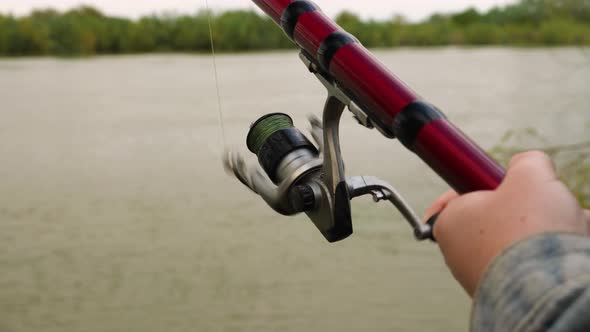 Close-up of a woman's hand spinning the handles with the reel of a red fishing rod against a river alt