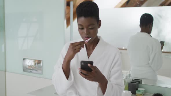 African american attractive woman brushing teeth and using smartphone in bathroom alt