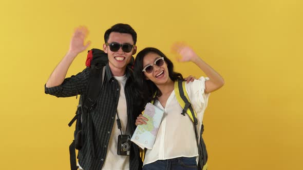 Happy Asian couple tourist backpackers waving hands and smiling on yellow stuio background