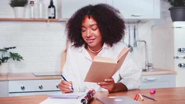 Young African American Woman Makes Notes in Workbook Sits at Kitchen alt