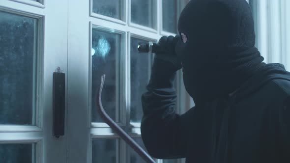 Close Up Of Thief Man Holding Flashlight And Crowbar Looking Inside The House While Standing Outside alt