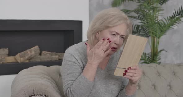 Elegant Senior Woman Examining Wrinkles on Face Using Mirror. Portrait of Worried Middle-aged Female alt