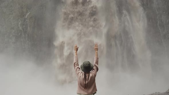 Woman traveler in hat standing with raised hands near big Karakaya-Su waterfall, powerful water flow