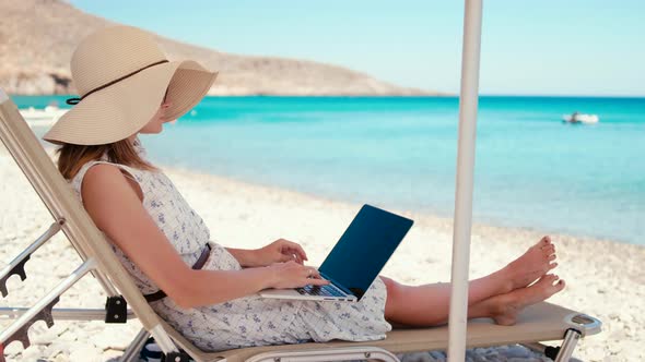 Beautiful Woman in Hat is Using Laptop and Relaxing on Beach By Sea on Vacation