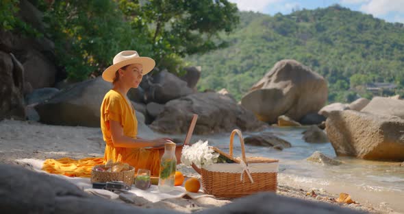 Young Woman in Hat and Yellow Dress Typing on Laptop Sitting on Beach Perfect Summer Day Outdoors alt
