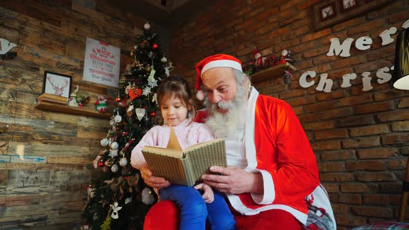 Santa Claus Sits on Armchair and Reads Book with Fairy Tales for Enthusiastic Little Girl in