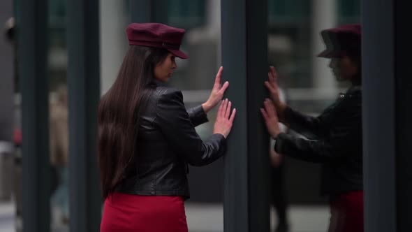 a Brunette with Long Hair in a Black Leather Jacket and a Burgundy Cap Stands Against a Glass Wall alt